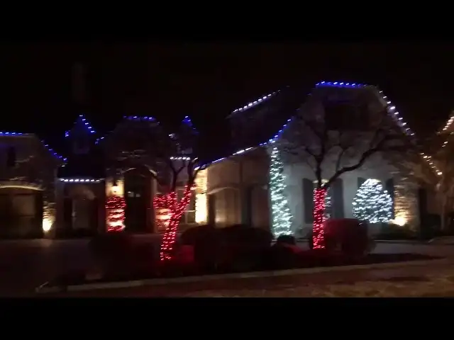 Home outlined in blue and white Christmas lights with lit trees at night
